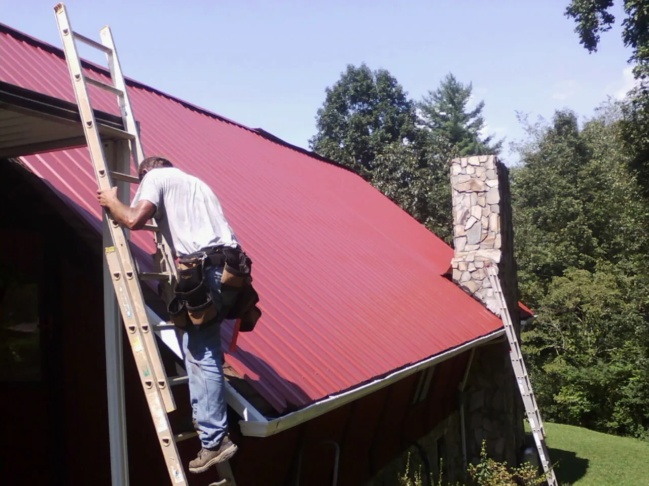 Roofer on a ladder beside a red metal roof and stone chimney — Soffit & Fascia Repair by Roofing Brothers in Bunn